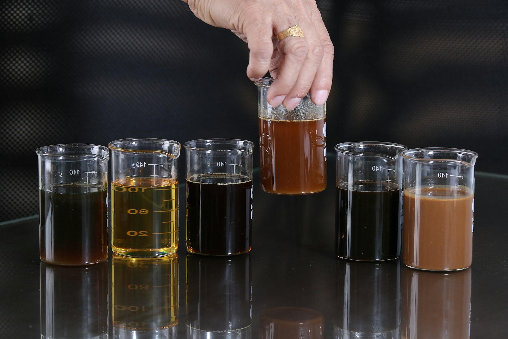 Six laboratory beakers on a reflective black bench holding liquids from pale gold to near-black, with a hand lifting the amber one out of the row