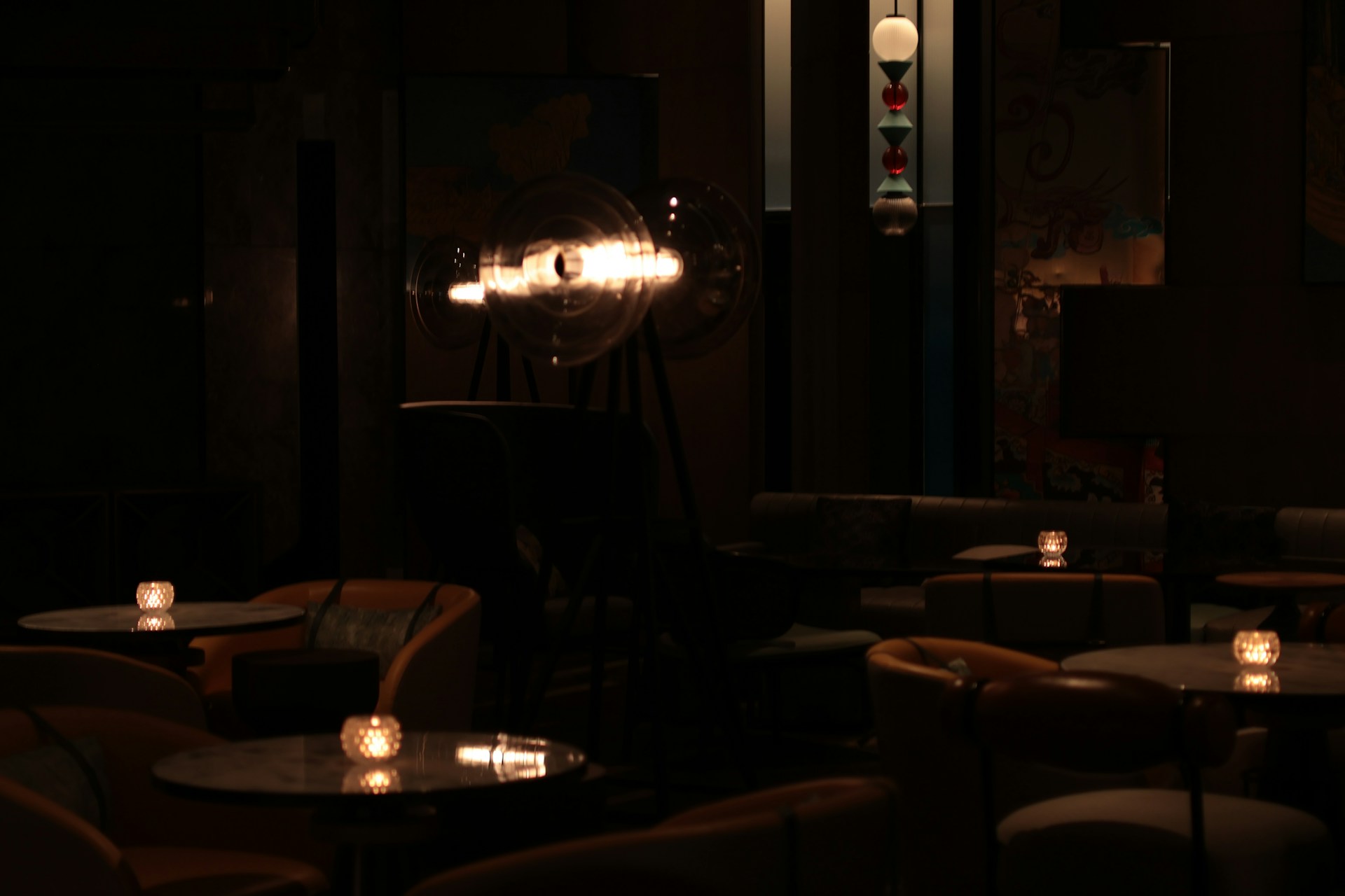 A dim restaurant room after service: marble tables with tealights, tan leather armchairs, and one large glass floor lamp burning in the middle distance