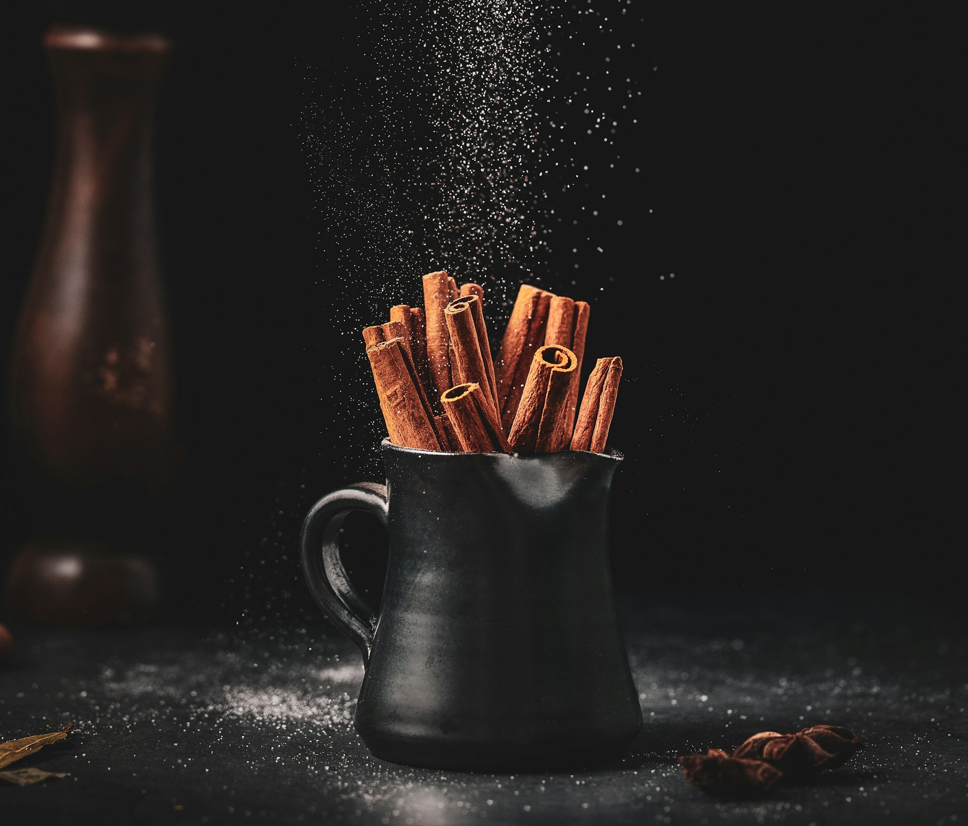 Cinnamon quills standing in a small black ceramic jug, with powder falling through the light above them against a black ground