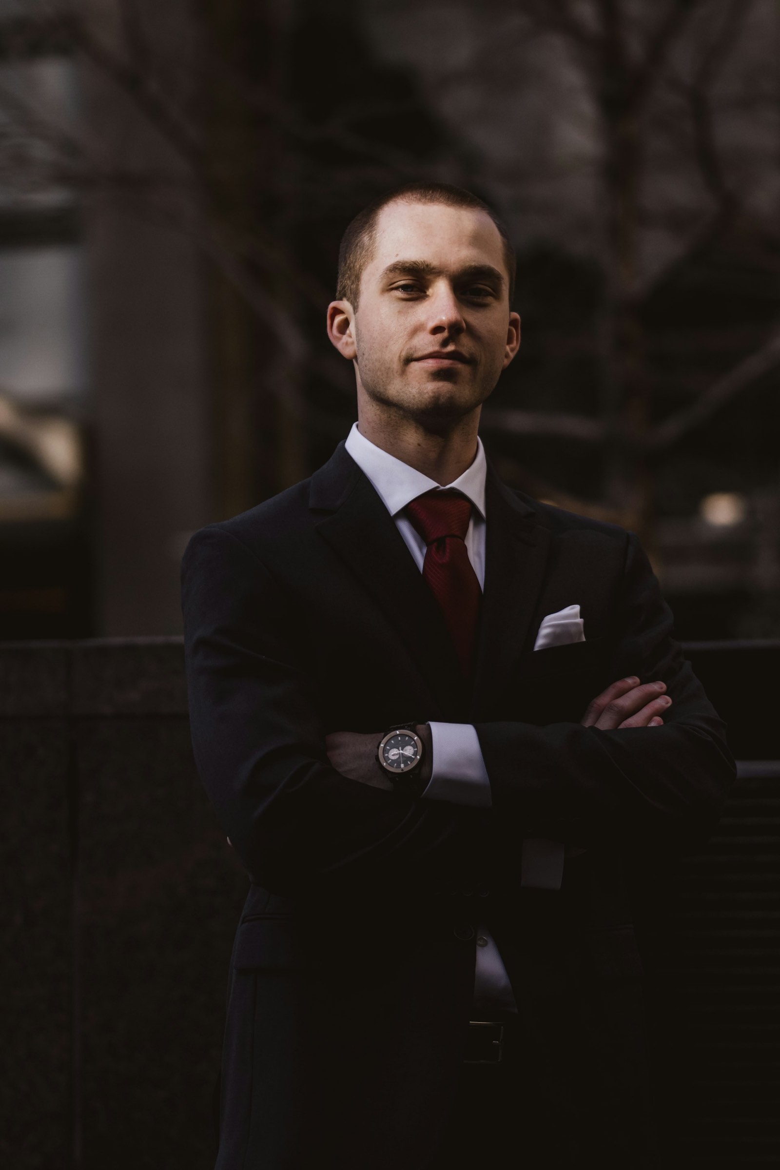 a young man in a dark suit with a red tie and folded arms, standing in front of a city building at dusk