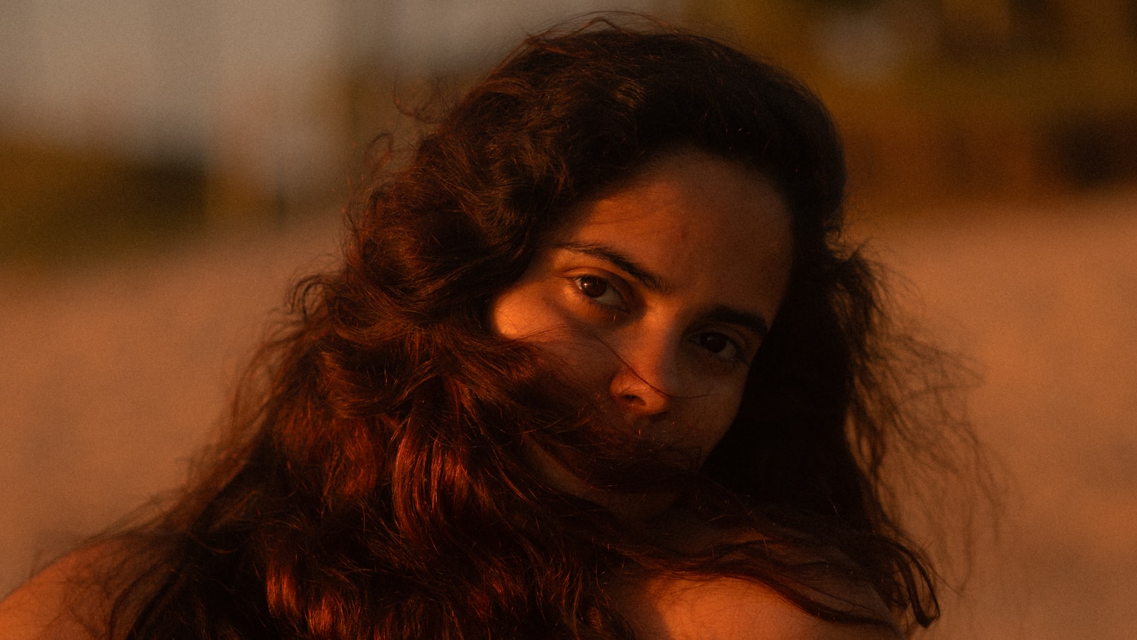 a woman with long dark curls blown across her face, looking back over her shoulder in warm low sunlight