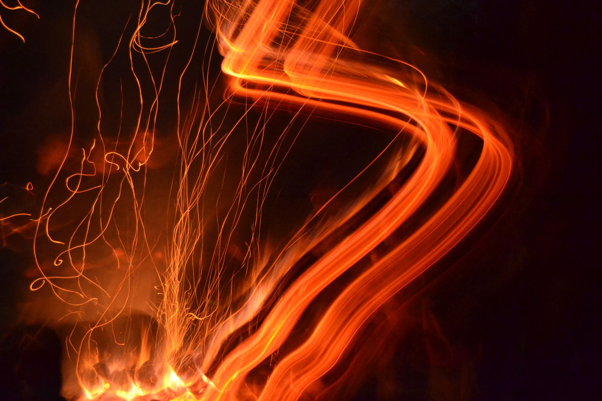 A long exposure of a fire at night: orange light trails sweeping in a bright curve across the frame with fine sparks trailing away to the left
