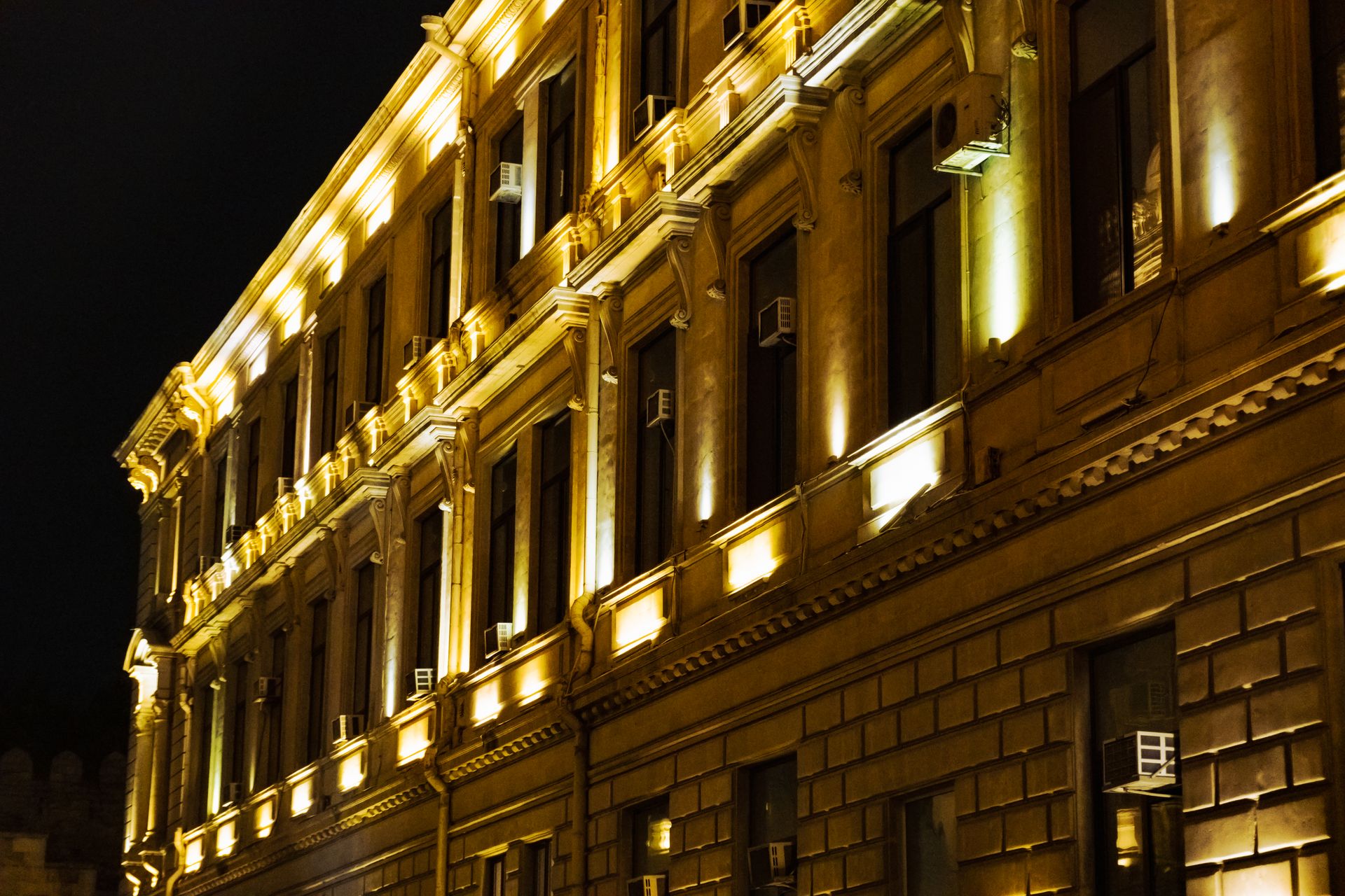 The upper floors of an ornate stone institutional building at night, washed with gold uplighting, its tall windows dark