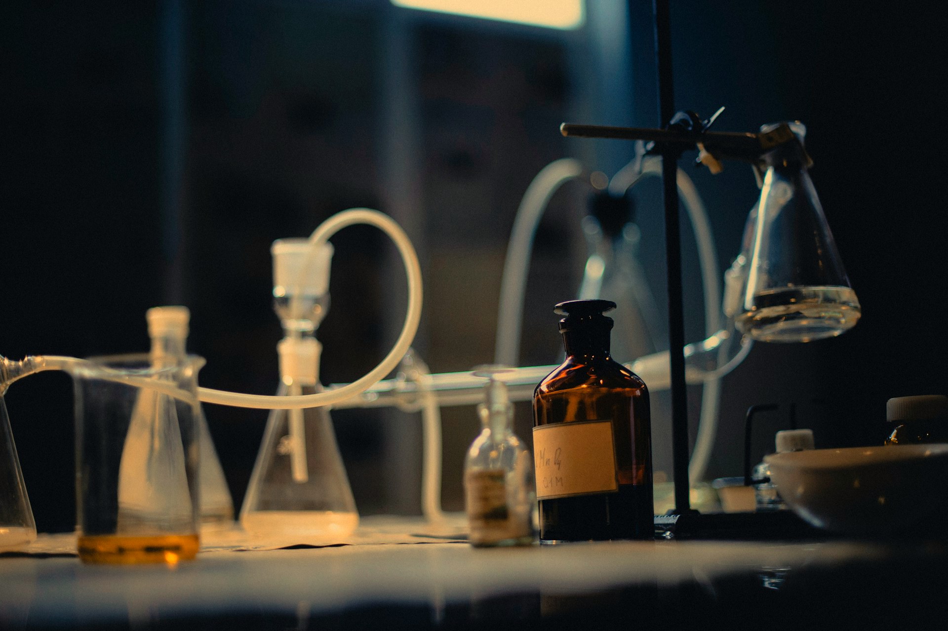 A dark laboratory bench at night: conical flasks and a beaker of amber liquid catching low light, with a brown reagent bottle and a clamp stand standing behind them