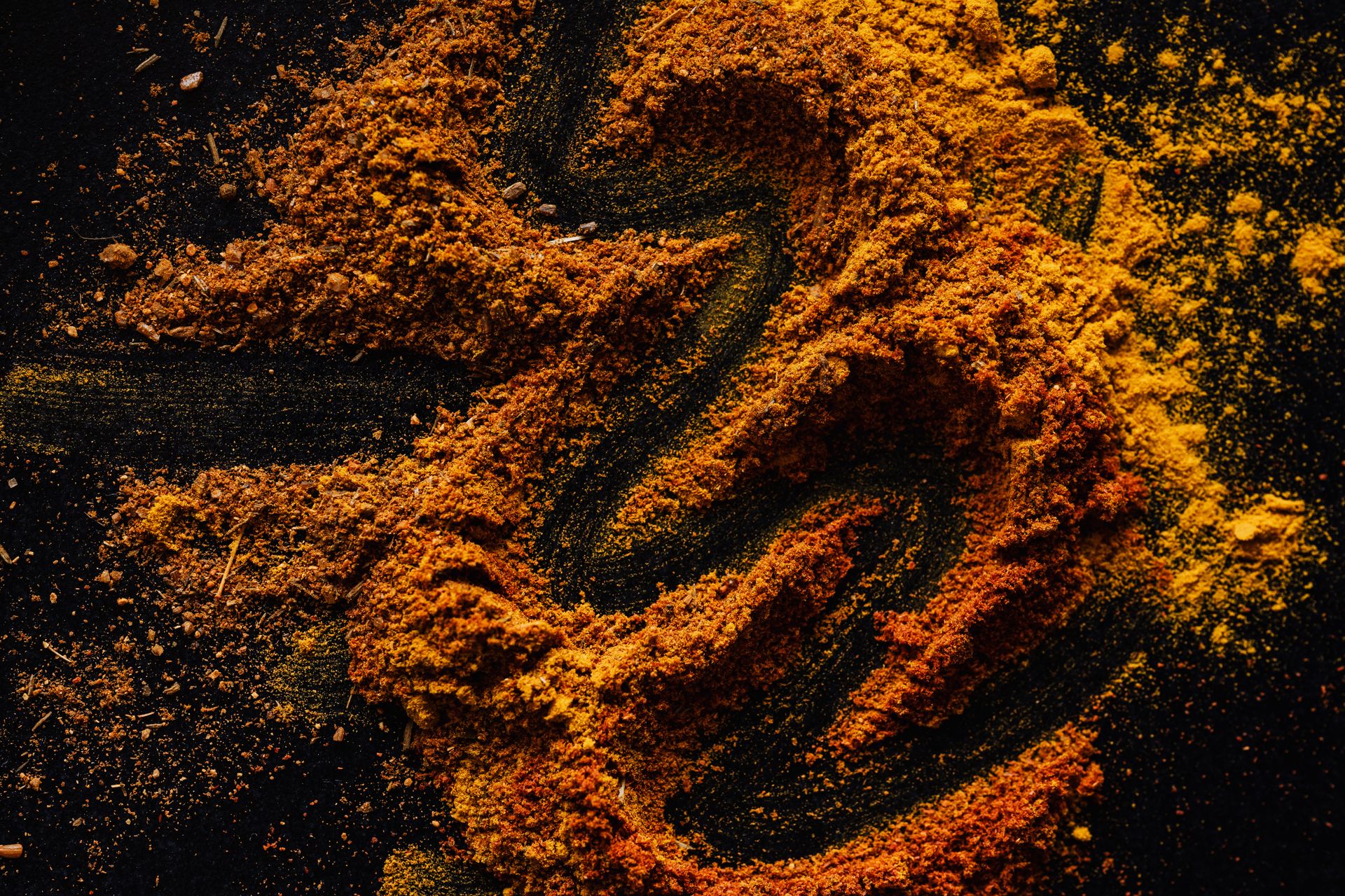 Milled orange and ochre spice powders swept across a black surface in loose arcs, coarse grains catching the light at the edges