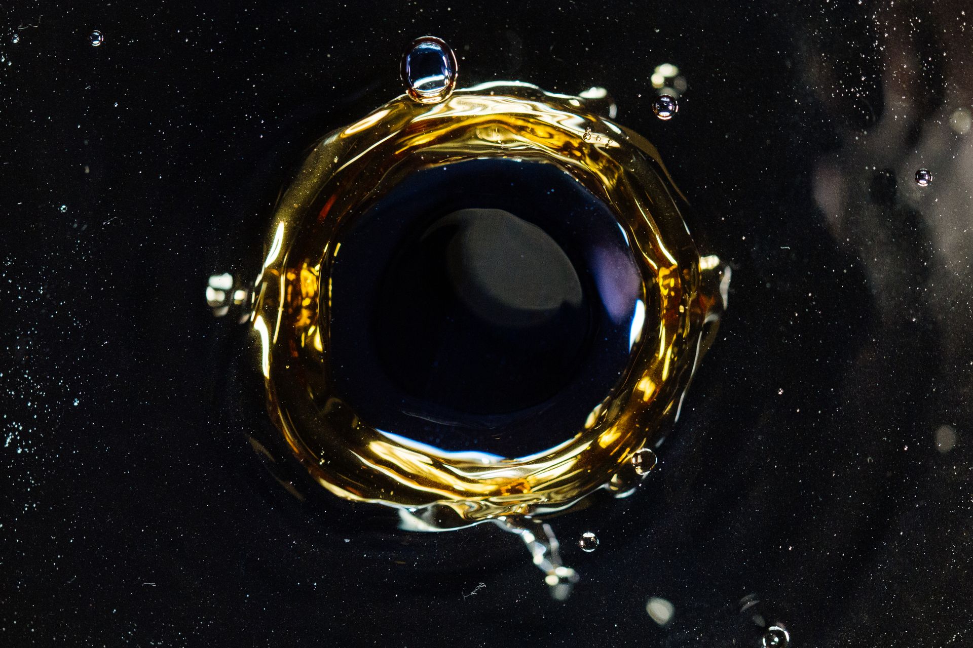 A macro photograph of a ring of golden oil floating on a dark liquid, forming a bright annulus around a near-black centre