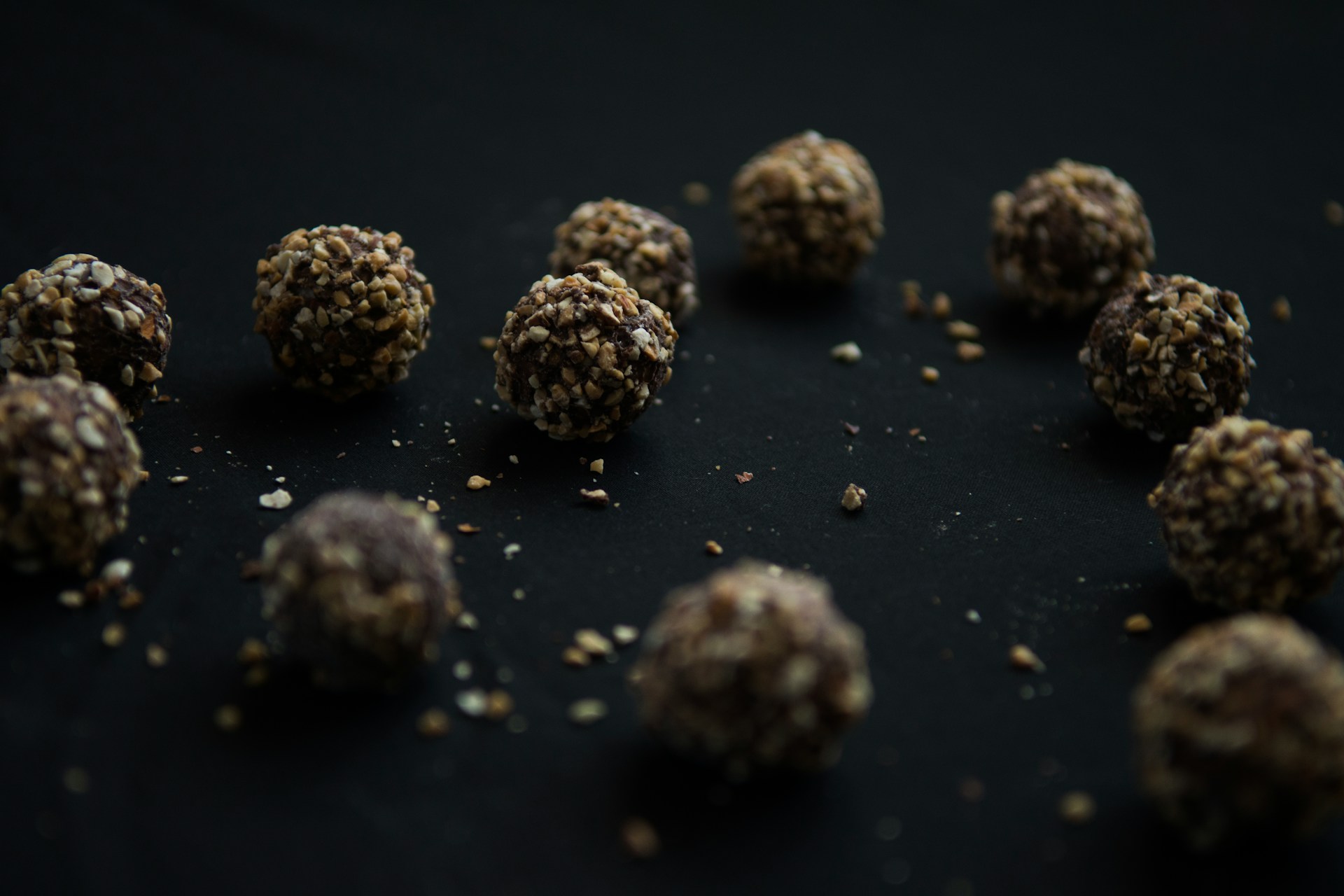 A dozen dark, roughly spherical units coated in chopped nuts and grain, arranged loosely on black cloth with crumbs fallen between them